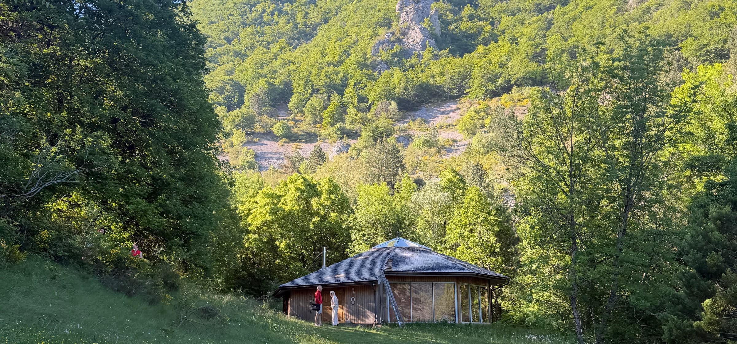 Une yourte en bois dans une prairie, au pied d'une montagne verdoyante, en plein soleil